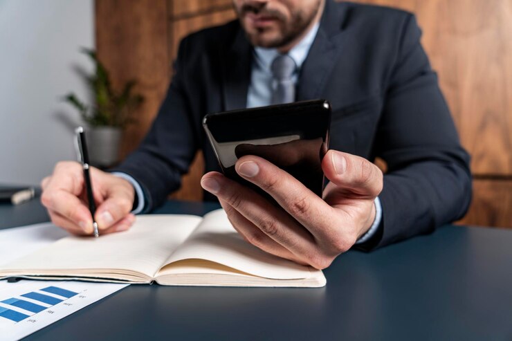 A businessman in a suit is sitting at a desk, writing in a notebook with a pen in one hand, while holding a smartphone in the other. A businessman in a suit is sitting at a desk, writing in a notebook with a pen in one hand, while holding a smartphone in the other.