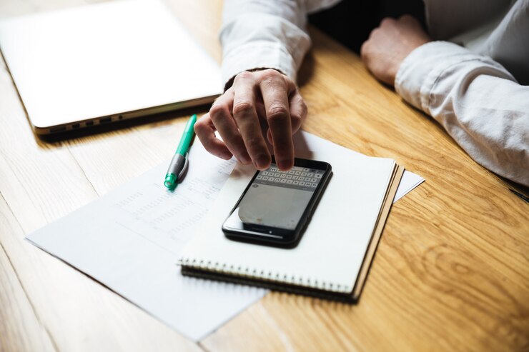 A person's hand is typing on a smartphone, with a laptop and paperwork on a table beside them. A person's hand is typing on a smartphone, with a laptop and paperwork on a table beside them.