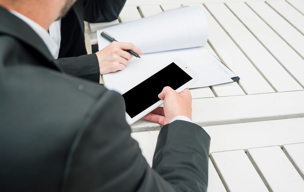 Two people, both wearing business attire, are sitting at a table. One person is reviewing documents and signing them with a pen, while the other is looking at a tablet. Two people, both wearing business attire, are sitting at a table. One person is reviewing documents and signing them with a pen, while the other is looking at a tablet.