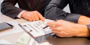 Two businessmen are sitting at a table, reviewing a document together. One man is pointing at a specific section of the document, while the other is holding a pen and listening attentively.