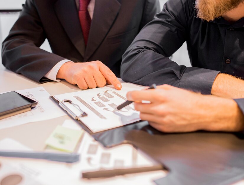Two businessmen are sitting at a table, reviewing a document together. One man is pointing at a specific section of the document, while the other is holding a pen and listening attentively. Two businessmen are sitting at a table, reviewing a document together. One man is pointing at a specific section of the document, while the other is holding a pen and listening attentively.