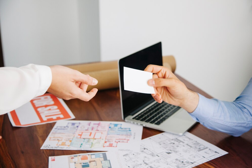 A real estate agent is handing a business card to a potential client. They are sitting at a table with a laptop, floor plans, and a "For Sale" sign. A real estate agent is handing a business card to a potential client. They are sitting at a table with a laptop, floor plans, and a "For Sale" sign.