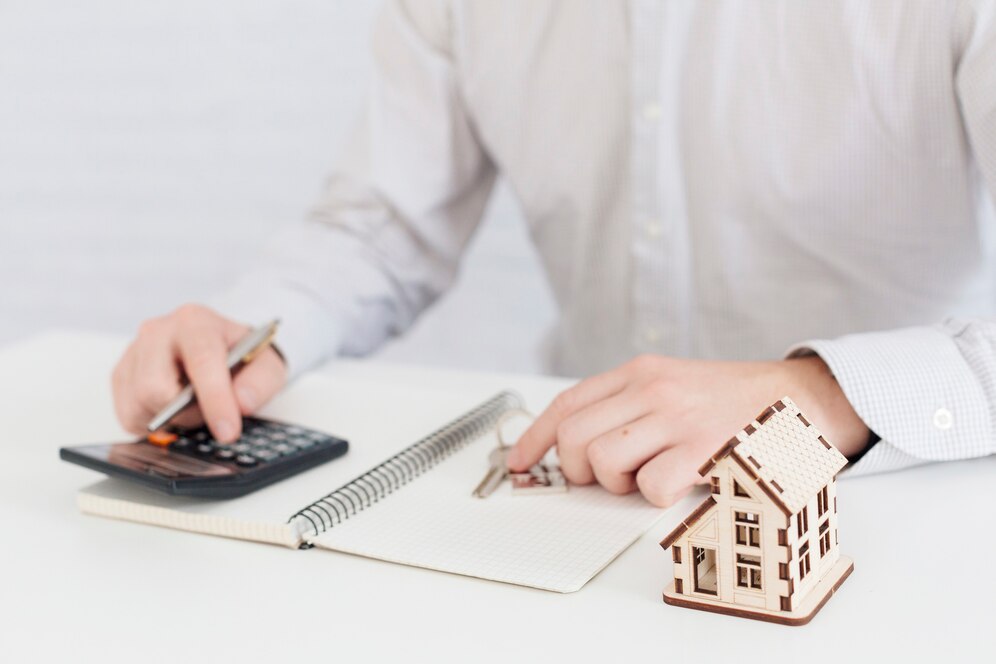 A person is sitting at a desk, using a calculator and a pen to make calculations. There is a notebook, house keys, and a small wooden model house on the desk. A person is sitting at a desk, using a calculator and a pen to make calculations. There is a notebook, house keys, and a small wooden model house on the desk.