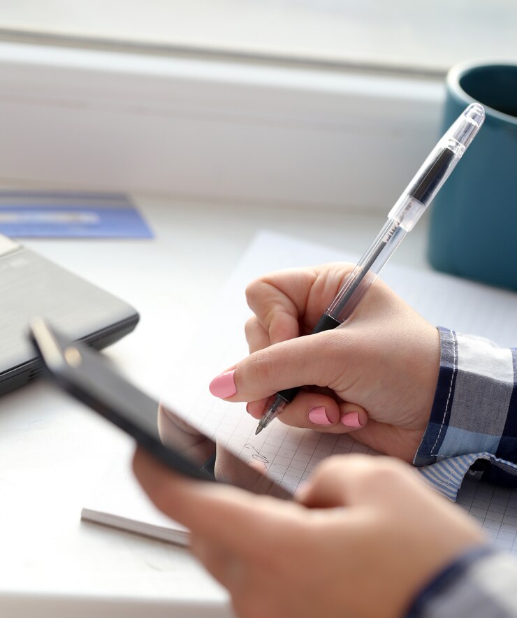 A woman's hands are shown writing in a notebook with a pen. She is also holding a smartphone in her other hand. A woman's hands are shown writing in a notebook with a pen. She is also holding a smartphone in her other hand.